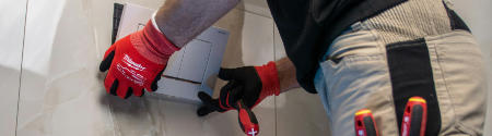 Eine Person mit roten Handschuhen und einer Arbeitshose installiert oder justiert einen an der Wand befestigten Elektrokasten mit einem Schraubenzieher, wobei das Werkzeug in der Gesäßtasche sichtbar ist - ein typisches Beispiel für Hausmeisterdienste.