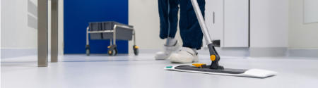 Eine Person mit weißen Schuhen wischt einen sauberen, hellen Boden in einem Flur, im Hintergrund steht ein Reinigungswagen - ein Zeichen für professionelles Facility Management und zuverlässige Hausmeisterdienste.